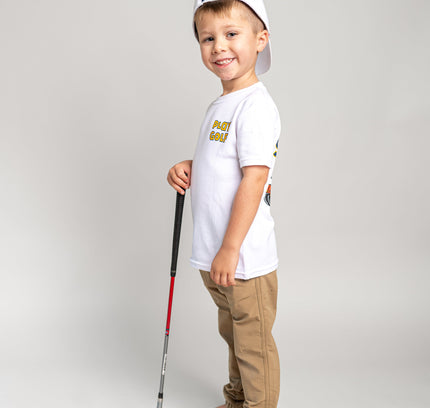 A young boy stands barefoot on a light gray studio background, wearing a white t-shirt with a yellow graphic, tan pants, and a white baseball cap backwards while holding a toy golf club.