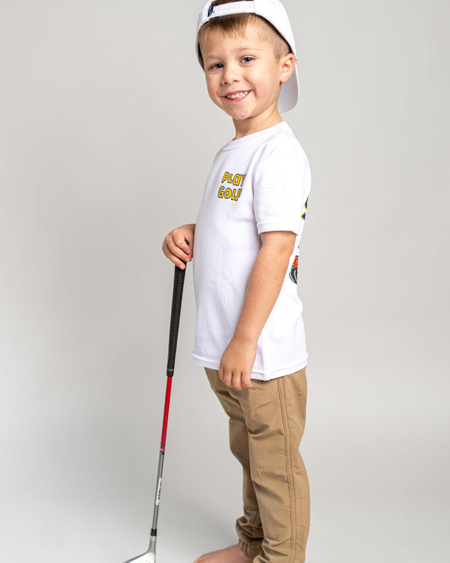 A young boy stands barefoot on a light gray studio background, wearing a white t-shirt with a yellow graphic, tan pants, and a white baseball cap backwards while holding a toy golf club.