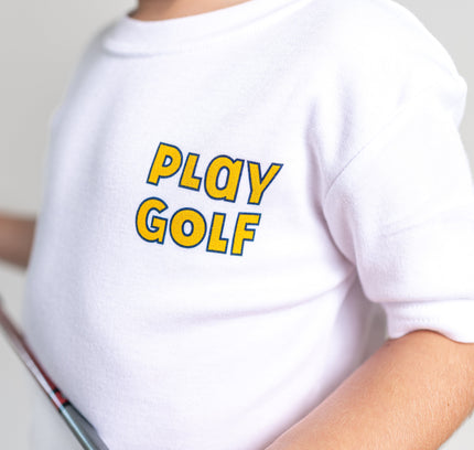 A close-up of a child wearing a white short-sleeve t-shirt with 'PLAY GOLF' printed in yellow and blue bubble letters on the chest, holding a golf club against a light gray background.