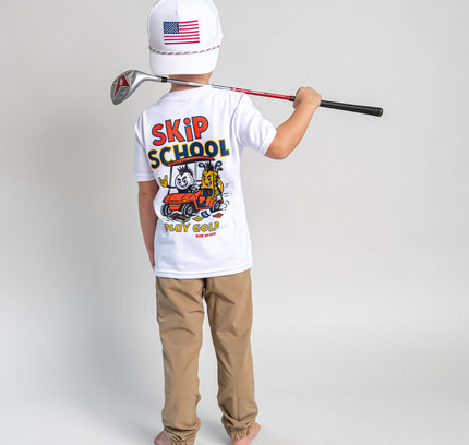 A child stands with their back to the camera wearing a white t-shirt with a colorful 'SKIP SCHOOL GOLF' graphic, khaki pants, and a white baseball cap with an American flag. They are holding a golf club over their shoulder against a light gray studio background.