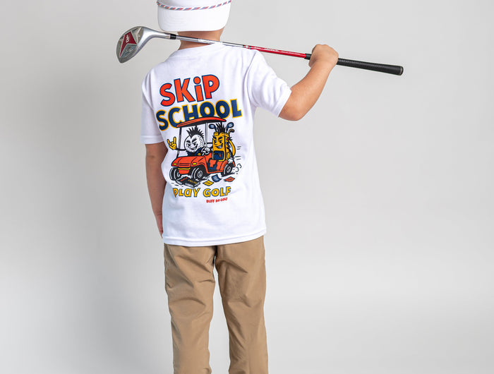 A child stands with their back to the camera wearing a white t-shirt with a colorful 'SKIP SCHOOL GOLF' graphic, khaki pants, and a white baseball cap with an American flag. They are holding a golf club over their shoulder against a light gray studio background.