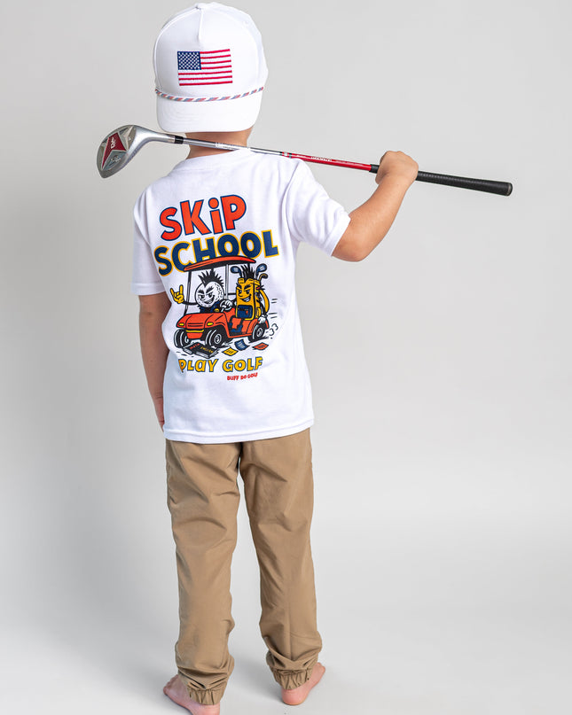 A child stands with their back to the camera wearing a white t-shirt with a colorful 'SKIP SCHOOL GOLF' graphic, khaki pants, and a white baseball cap with an American flag. They are holding a golf club over their shoulder against a light gray studio background.
