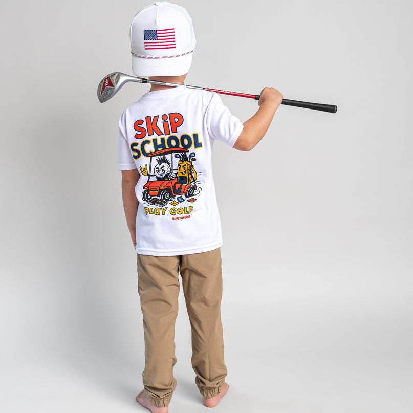 A child stands with their back to the camera wearing a white t-shirt with a colorful 'SKIP SCHOOL GOLF' graphic, khaki pants, and a white baseball cap with an American flag. They are holding a golf club over their shoulder against a light gray studio background.