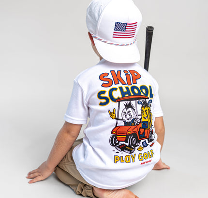 A child sitting on a light gray studio floor, shown from the back, wearing a white t-shirt with a colorful 'SKIP SCHOOL PLAY GOLF' graphic on the back, a white baseball cap with an American flag patch, and khaki pants.