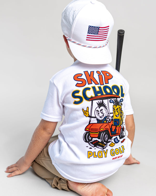 A child sitting on a light gray studio floor, shown from the back, wearing a white t-shirt with a colorful 'SKIP SCHOOL PLAY GOLF' graphic on the back, a white baseball cap with an American flag patch, and khaki pants.