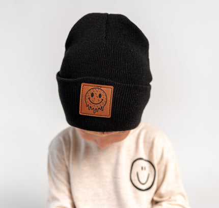 A child wearing a black knit beanie with a brown leather patch featuring a smiley face and the brand name 'Sodapop', paired with a cream sweatshirt against a plain light gray background.
