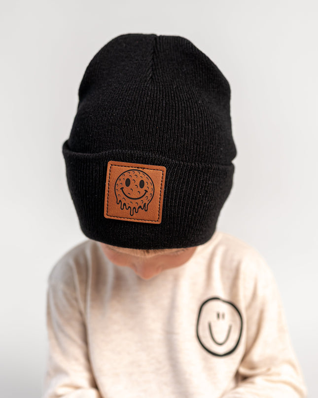 A child wearing a black knit beanie with a brown leather patch featuring a smiley face and the brand name 'Sodapop', paired with a cream sweatshirt against a plain light gray background.