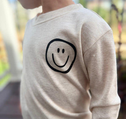 A close-up side view of a child wearing a cream-colored long-sleeve shirt with a simple black smiley face graphic on the chest, set against a blurred outdoor background.