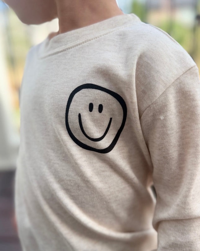 A close-up side view of a child wearing a cream-colored long-sleeve shirt with a simple black smiley face graphic on the chest, set against a blurred outdoor background.