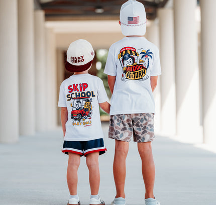 Two children standing from behind in a covered walkway, wearing matching white graphic T-shirts and shorts. The child on the left wears navy shorts with red trim and white sneakers, and the child on the right wears camouflage shorts and white sneakers.