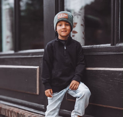 A young boy wearing a black quarter-zip pullover, light-wash jeans, tan boots, and a gray beanie with a red logo, sitting on the brick ledge of a dark building.