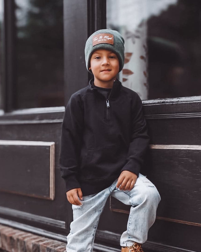 A young boy wearing a black quarter-zip pullover, light-wash jeans, tan boots, and a gray beanie with a red logo, sitting on the brick ledge of a dark building.