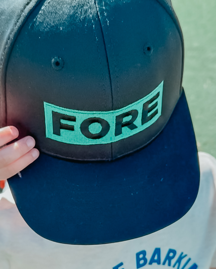 Child wearing a black golf hat with a bold green embroidered patch reading 'FORE,' playful and stylish golf accessory for kids, photographed outdoors on a sunny day.