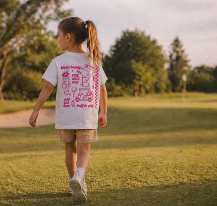 Child walking on a grassy field wearing a white t-shirt with pink graphics