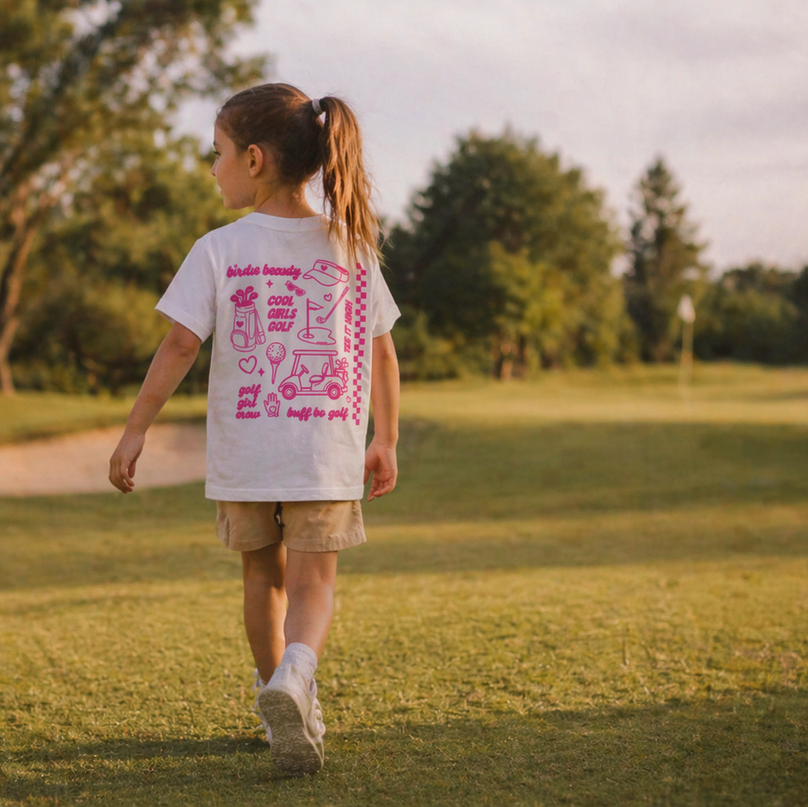 Child walking on a grassy field wearing a white t-shirt with pink graphics
