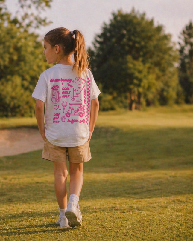 Young girl walking on a golf course wearing a white t-shirt with pink text and graphics.
