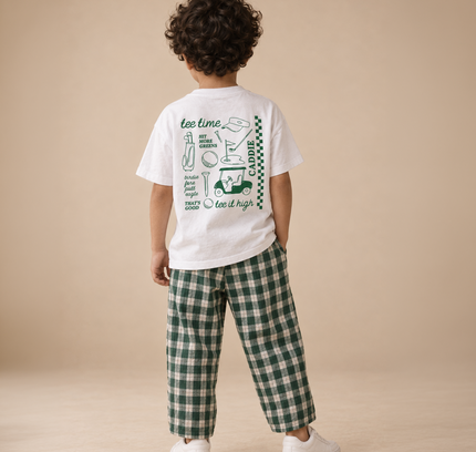 A child standing with their back to the camera, wearing a white graphic t-shirt with green illustrations and green-and-white plaid pants, on a beige studio background.
