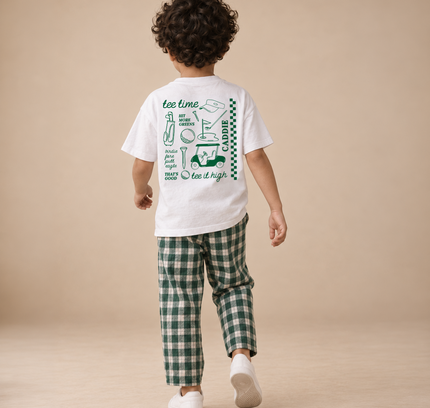 A child shown from behind wearing a white graphic t-shirt and green plaid pants, walking on a neutral beige studio floor.