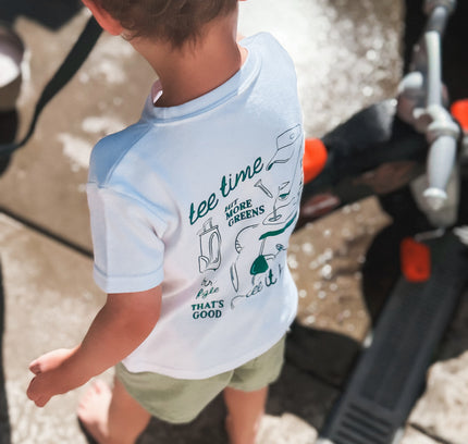 Child wearing a light blue and white t-shirt with text and graphics, standing on a sidewalk.
