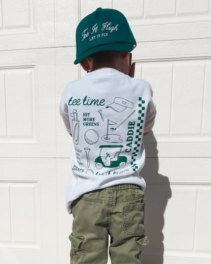 Child wearing a white t-shirt with graphics and text, green pants, and a green cap in front of a white garage door.