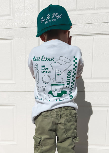 Child wearing a white t-shirt with graphics and text, green pants, and a green cap in front of a white garage door.