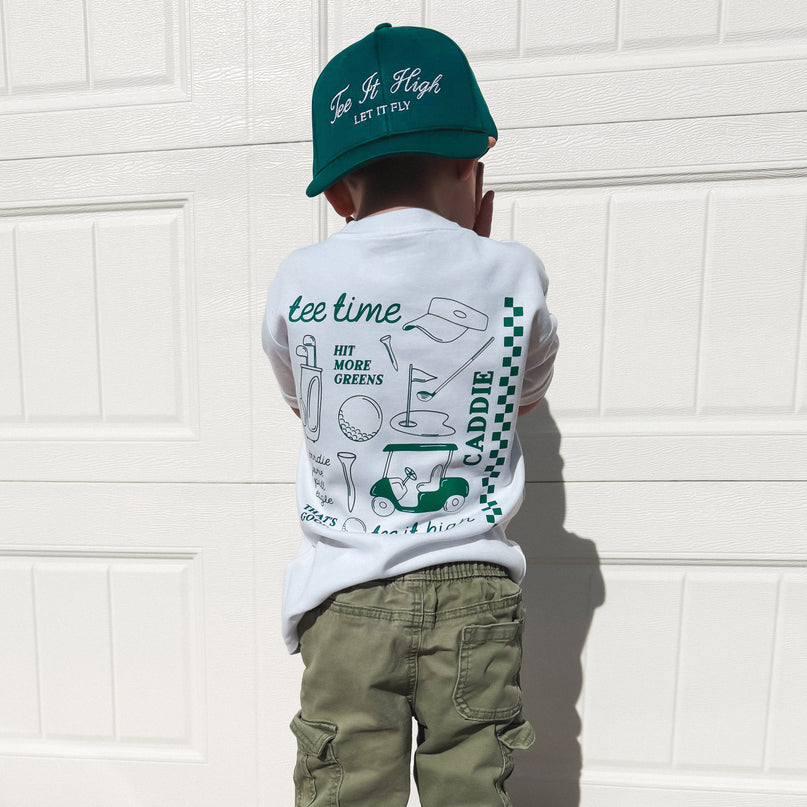 Child wearing a white t-shirt with graphics and text, green pants, and a green cap in front of a white garage door.