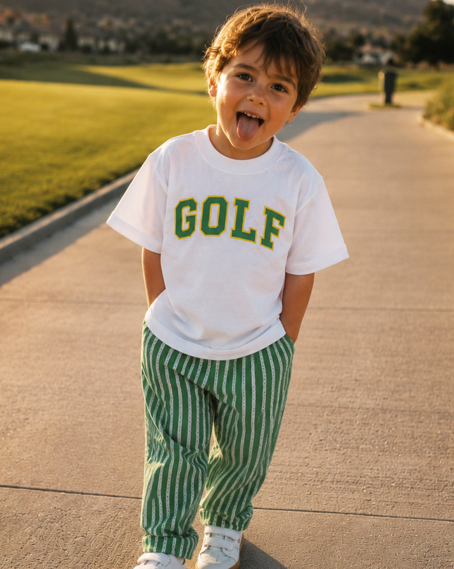 Child wearing a 'GOLF' shirt and green striped pants standing on a path with mountains in the background.