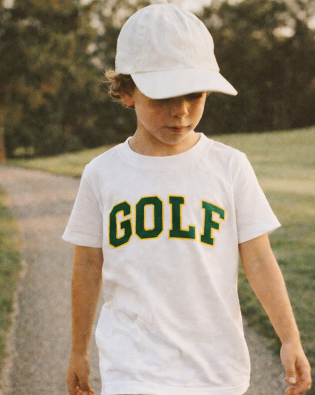 Child wearing a white t-shirt with 'GOLF' text, beige shorts, and a white cap walking on a path.