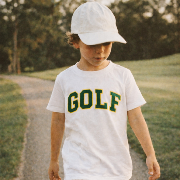 Child wearing a white t-shirt with 'GOLF' text, beige shorts, and a white cap walking on a path.