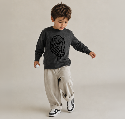 A toddler boy wearing a dark gray long-sleeve crewneck sweatshirt with a black astronaut graphic, paired with beige cargo pants and black-and-white sneakers, walking against a neutral beige studio background.