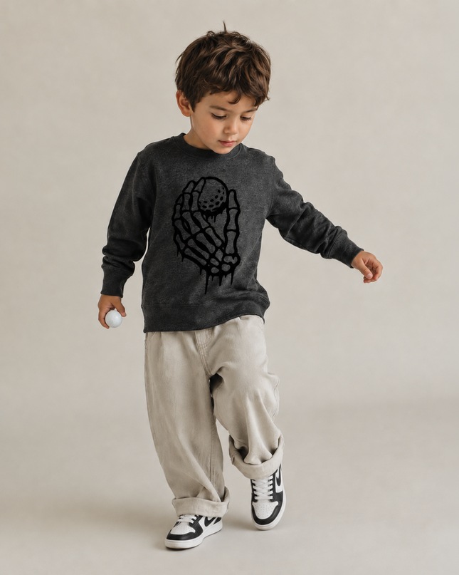 A toddler boy wearing a dark gray long-sleeve crewneck sweatshirt with a black astronaut graphic, paired with beige cargo pants and black-and-white sneakers, walking against a neutral beige studio background.