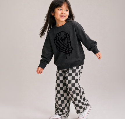 A young girl smiling and posing in a dark charcoal gray sweatshirt with a black skeleton hand graphic, paired with black and white checkered pants and white sneakers, against a solid light gray studio background.