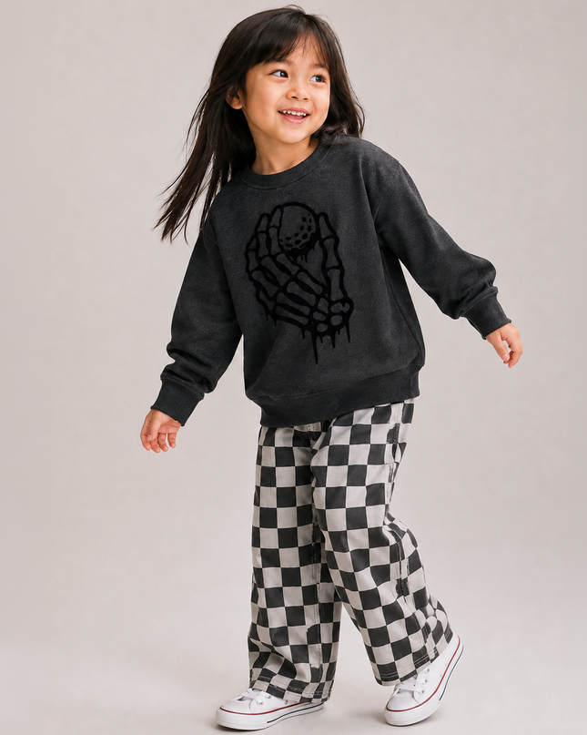 A young girl smiling and posing in a dark charcoal gray sweatshirt with a black skeleton hand graphic, paired with black and white checkered pants and white sneakers, against a solid light gray studio background.