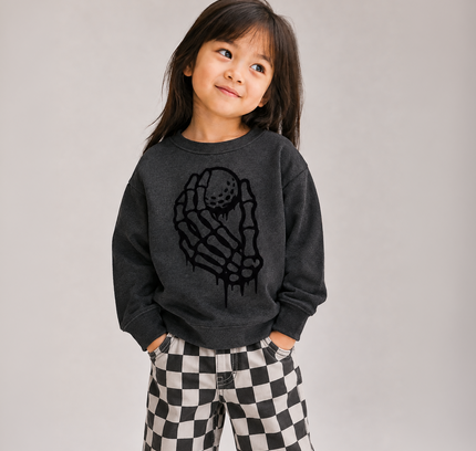 A young girl with dark hair is wearing a dark gray sweatshirt with a black dripping skeleton hand graphic and black-and-white checkered pants, standing against a neutral light gray background.