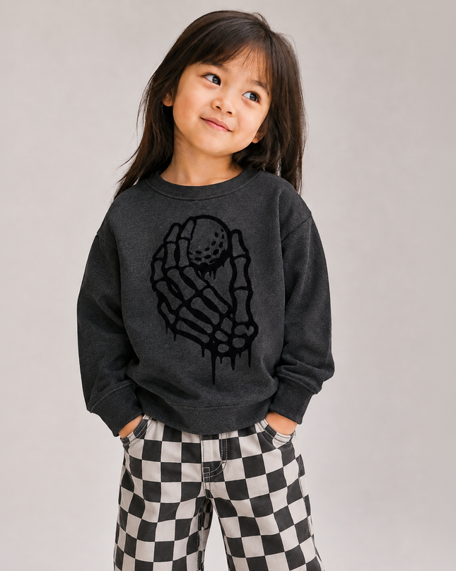 A young girl with dark hair is wearing a dark gray sweatshirt with a black dripping skeleton hand graphic and black-and-white checkered pants, standing against a neutral light gray background.