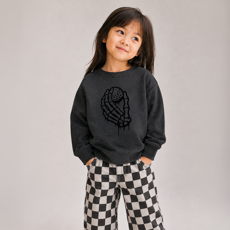 A young girl with dark hair is wearing a dark gray sweatshirt with a black dripping skeleton hand graphic and black-and-white checkered pants, standing against a plain light gray background.