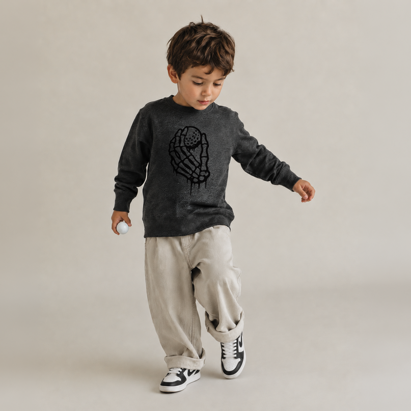 A toddler boy wearing a dark gray long-sleeve crewneck sweatshirt with a black astronaut graphic, paired with beige cargo pants and black-and-white sneakers, walking against a neutral beige studio background.
