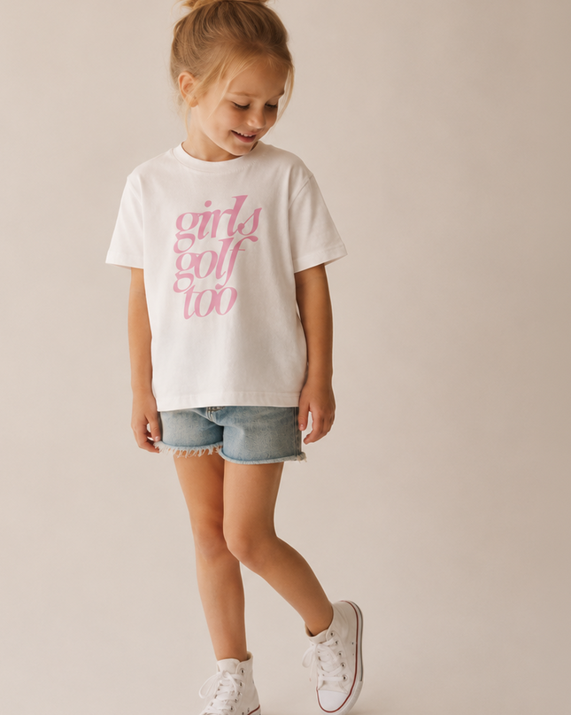 A young girl with her hair in a bun is wearing a white t-shirt with pink text that reads 'girls golf too', paired with denim shorts and white sneakers, standing against a solid beige studio background.