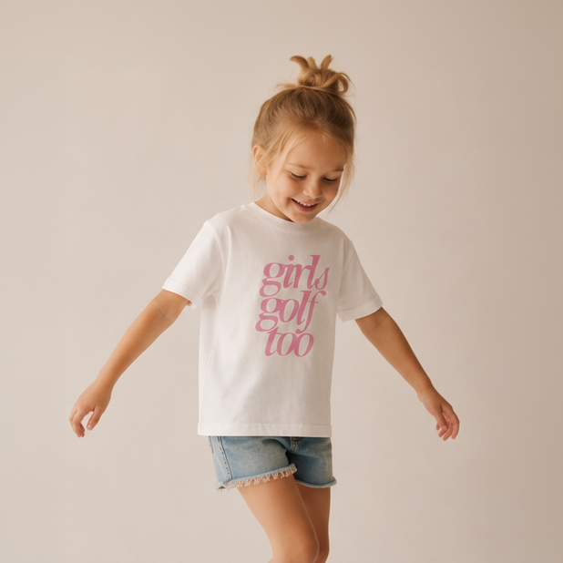 A young girl with her hair in a messy bun is wearing a white t-shirt with pink text that reads 'girls golf too' and denim shorts, posing against a solid light beige background.