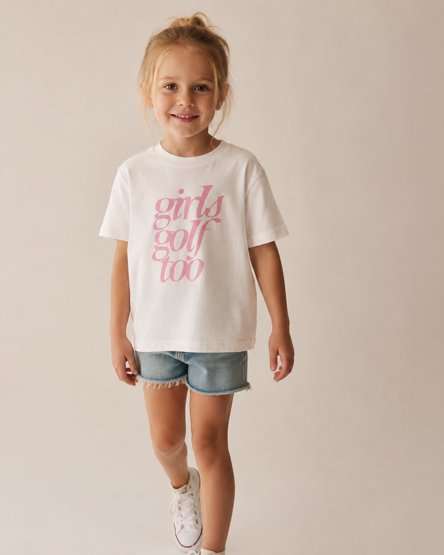 A young girl with blonde hair tied up in a bun is wearing a white short-sleeve t-shirt with pink text that reads 'girls golf too', paired with denim shorts and white sneakers, standing against a plain beige studio background.