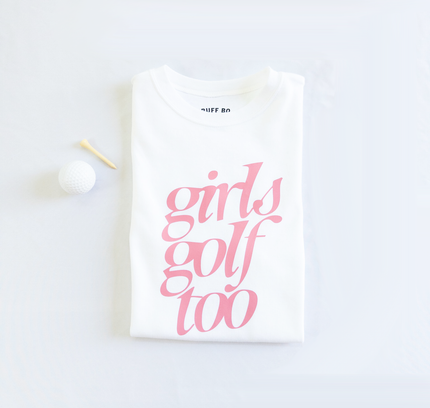 Folded white t-shirt with pink text that reads 'girls golf too,' placed next to a white golf ball and wooden tee on a light background.