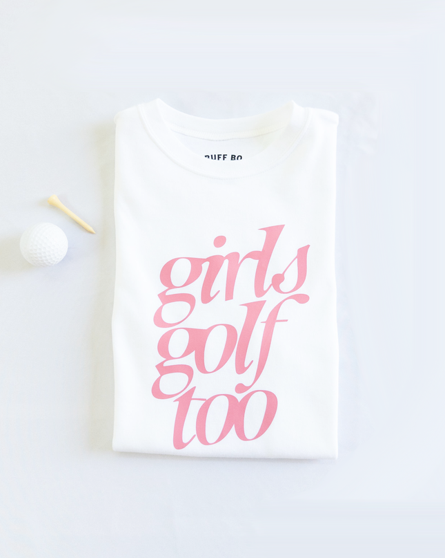 Folded white t-shirt with pink text that reads 'girls golf too,' placed next to a white golf ball and wooden tee on a light background.
