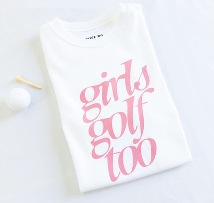 Folded white t-shirt with pink text that reads 'girls golf too,' placed next to a white golf ball and wooden tee on a light background.