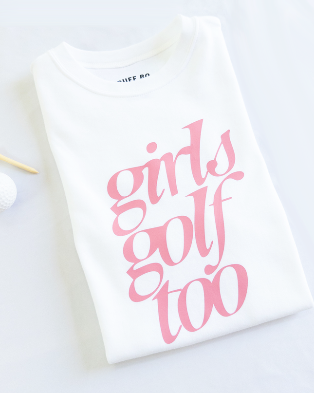 Folded white t-shirt with pink text that reads 'girls golf too,' placed next to a white golf ball and wooden tee on a light background.