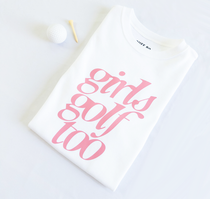 Folded white t-shirt with pink text that reads 'girls golf too,' placed next to a white golf ball and wooden tee on a light background.