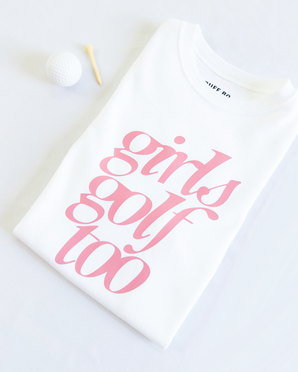 Folded white t-shirt with pink text that reads 'girls golf too,' placed next to a white golf ball and wooden tee on a light background.