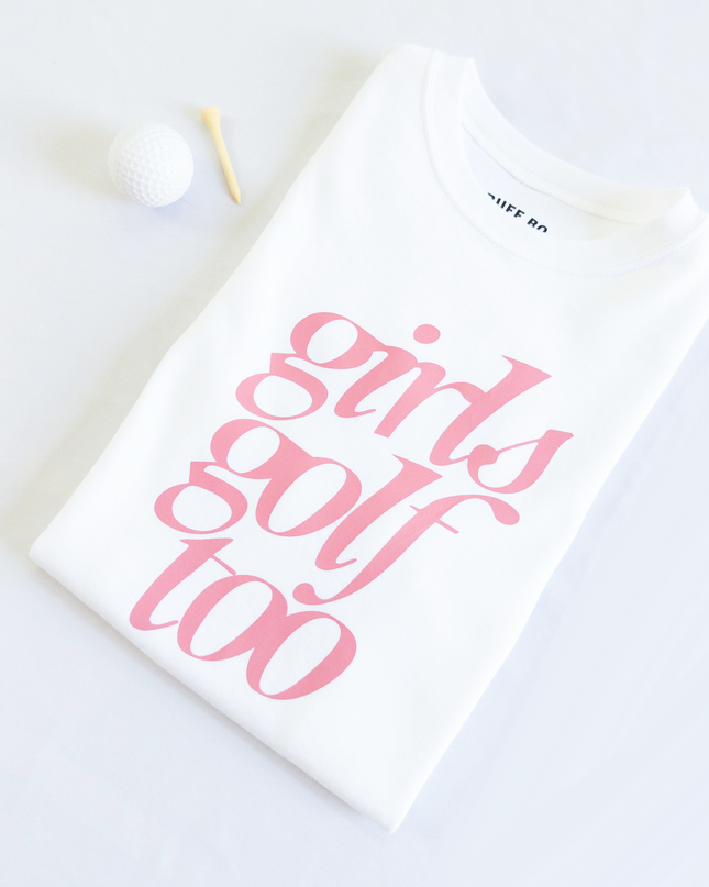 Folded white t-shirt with pink text that reads 'girls golf too,' placed next to a white golf ball and wooden tee on a light background.