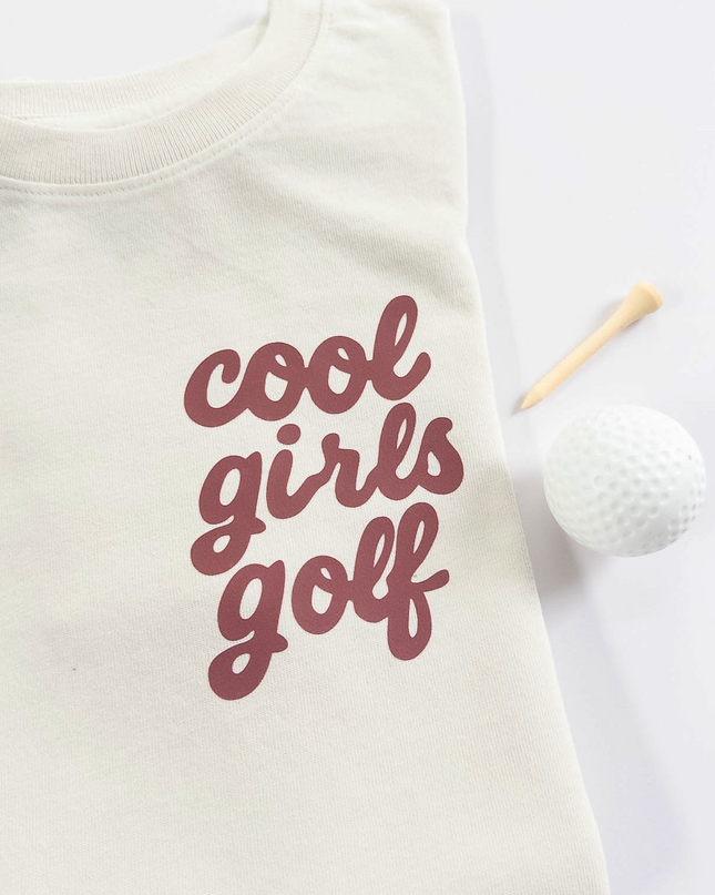 Folded white t-shirt with 'cool girls golf' text on a light background