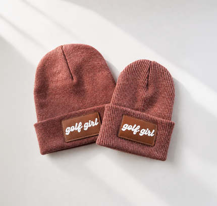 Two ribbed knit beanie hats in a dusty rose-brown color, laid flat on a light gray surface with soft diagonal shadows. Each beanie has a brown rectangular patch on the folded cuff with the words 'golf girl' in white script.