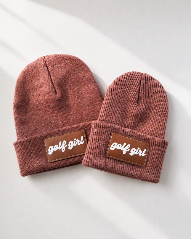 Two ribbed knit beanie hats in a dusty rose-brown color, laid flat on a light gray surface with soft diagonal shadows. Each beanie has a brown rectangular patch on the folded cuff with the words 'golf girl' in white script.